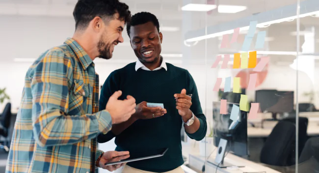 Two men smiling and discussing ideas, standing by an office whiteboard with colorful sticky notes.