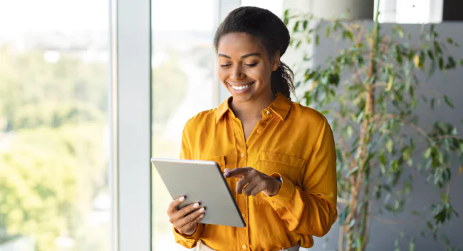 Smiling woman in yellow shirt using a tablet near a window and indoor plant.