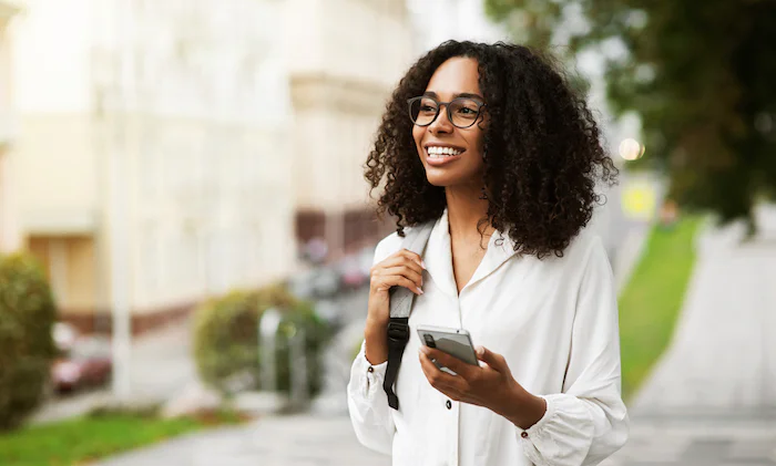 Woman with curly hair and glasses smiles while holding a smartphone and wearing a backpack.