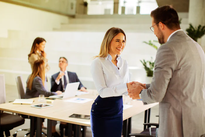 Woman in business attire shaking hands with a man in an office setting, with colleagues working in the background.